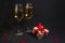 Two glasses of champagne stand in the dark, next to a gift box tied with a red ribbon, and confetti made of hearts