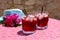 Two glasses of summer red cocktail with ice next to a blue hat and a sprig of Bougainvillea flowers on a pink table