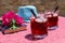 Two glasses of summer red cocktail with ice next to a book, a sprig of Bougainvillea flowers, blue hat on a pink table