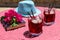 Two glasses of summer red cocktail with ice next to a book, a sprig of Bougainvillea flowers, blue hat on a pink table