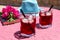 Two glasses of summer red cocktail with ice next to a book, a sprig of Bougainvillea flowers, blue hat on a pink table