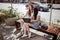 Two good looking thin girls with long hair,dressed in casual style, sit on the bench and hold a laptop on their knees.