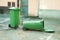 Two green plastic garbage can one is overturned lying on the street.