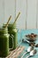 Two green smoothies in mason jar mugs with straws on a table wit