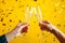 two hands clinking champagne glasses against vibrant yellow background with gold confetti