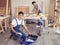 Two handsome Asian male repairmen or carpenters working together in wood workshop, one man sitting smiling and looking at camera