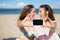 Two happy girls sitting on beach taking selfie laughing