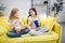 Two happy and joyful teenagers sit on yellow sofa. They look at each other and smile. Girls have bowl with popcorn and
