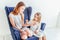 Two happy kids sitting on cozy blue chair relaxing playing in white living room indoors. Little girl playing with teenage girl