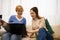 Two happy pretty women leafing through a catalog, laughing and talking while sitting on a comfortable sofa while waiting for a