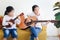 Two happy sister girls singing a song and playing guitar ukulele while sitting on sofa in living room, children spending special