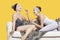 Two happy young women applying face pack while sitting on sofa over yellow background