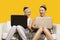 Two happy young women using laptop sitting on sofa against yellow background