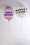 two helium balloons decorated with colored dots and a happy birthday message, with a hanging ribbon, on the roof of a white room,