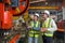 Two industrial workers engrossed in a discussion beside a high-tech assembly line machine, inside a bustling factory. Safety gear