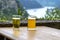 Two large mug of beer standing on a wooden table in the background a blurry river and valley.