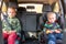 Two little boys sitting on a car seat and a booster seat buckled up in the car