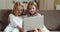 Two little girls sitting on couch looking at laptop, blonde sisters at home together watching movie or video online in