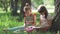 Two little girls under tree enjoying sunny day and reading book. older sister reads a book to her younger sister