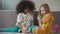 Two little multiracial girls sitting on couch and playing with teddy bears