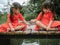 Two little sibling girls in red dress role playing fishing sitting on a wooden bridge by the pond. Childhood happiness