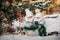 Two little twin girls in white suits take out Christmas balls from a basket near the Christmas tree in winter on the street