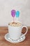 Two lollipops in shape of skull and coffin, sticking in cap of hot cocoa, chocolate or coffee latte with marshmallows on wooden ta
