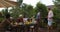 Two Man Taking Food From Barbecue People Sitting At Table Young Friends Group Gathering On Summer Terrace Having Party