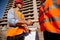 Two men dressed in shirts, orange work vests and helmets shake hands against the background of a multistorey building