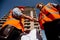 Two men dressed in shirts, orange work vests and helmets shake hands against the background of a multistorey building