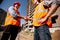 Two men dressed in shirts, orange work vests and helmets shake hands against the background of a multistorey building