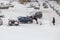 Two men pushing stuck Hyundai Solaris car through a snowy yard between rows of parked cars in deep snow in slope.