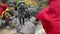 Two men in raincoats help a young woman with a backpack and trekking sticks to jump over a stormy river.