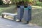 Two metal trash cans and a bench on a city street