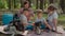 Two moms and two sons drinking tea on picnic blanket during summer family camping vacation with tent in forest. Boy