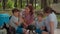 Two mothers and two sons drinking tea on picnic blanket during summer family camping vacation with tent in forest. Happy