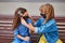 Two multi-ethnic adorable 10-year-old girls sit on a bench, one girl putting on another antiviral mask, outdoors.