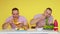 Two muscular twins eat healthy food and fast food at the table on a yellow background. The concept of healthy and