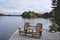 Two Muskoka chairs sitting on a wood dock facing a calm lake. Across the water is a cottage nestled among green trees