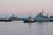 Two naval harbor tugs against the background of warships in the Petrovskaya pier of Kronstadt. Evening sunset. Russia