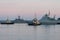 Two naval harbor tugs against the background of warships in the Petrovskaya pier of Kronstadt. Evening sunset. Russia