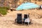 Two navy blue rusty beach chairs under a light blue striped umbrella on a beach.