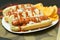 Two New York style hot dogs with ketchup, mustard, onions, and a side of potato chips, served on a white paper plate