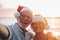 Two old happy seniors wearing christmas hats at the beach taking a selfie of them smiling and having fun with the sunset at the