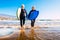 Two old and mature people having fun and enjoying their vacations outdoors at the beach wearing wetsuits and holding a surfboard