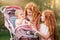 Two older redhead sisters are walking in the park on a sunny summer day with stroller with their younger sister. The girls try to