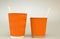 Two orange grooved paper cups for coffee stand on a white background, large and small with wooden spoons