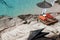 Two orange sun loungers under a straw beach umbrella on a rocky beach with azure water during the end of the holiday season