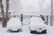 Two parked snow-covered cars in courtyard during plentiful snowfall.