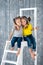 Two pretty cheerful girls sisters in jeans are sitting on a stepladder in front of a wooden wall background in studio